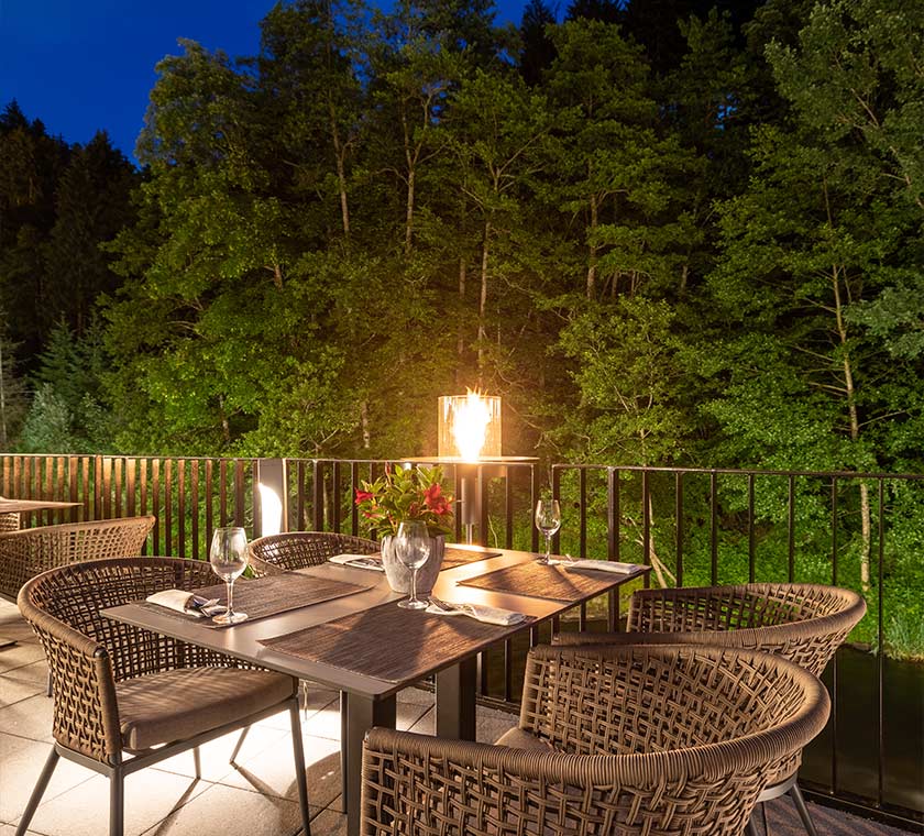 Abendstimmung mit Blick auf eine Tischgruppe auf unserer Murg-Terrasse. Im Hintergrund ist der an das Hotel angrenzende Wald zu erkennen. Im Vordergrund sieht man eine gedeckte Tischgruppe mit vier Plätzen. Die Stühle sind hellbraune Ratansessel, auf denen weiße Sitzkissen platziert wurden. Auf dem Tisch mit cremefarbener Tischplatte sind drei Gedecke vollständig und ein Gedeck versteckt hinter einer Tischpflanze zu sehen. Im rechten Hintergrund kann man hinterhalb der schwarzen Terrassenbegrenzung die Murg erkennen. Mittig im Hintergrund ist ein kleiner Tisch Kamin zu erkennen.